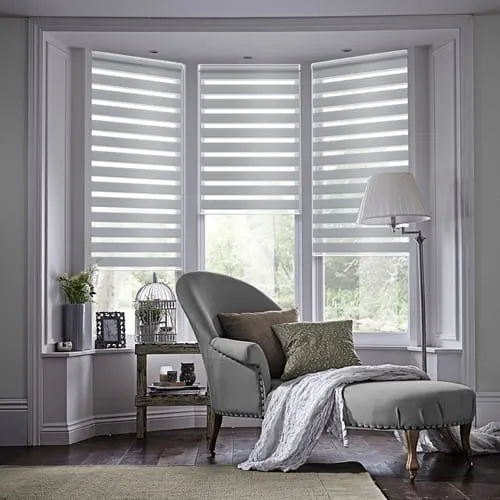 A grey chaise longue with neutral coloured furnishings against three tall windows styled with day and night blinds in Suffolk